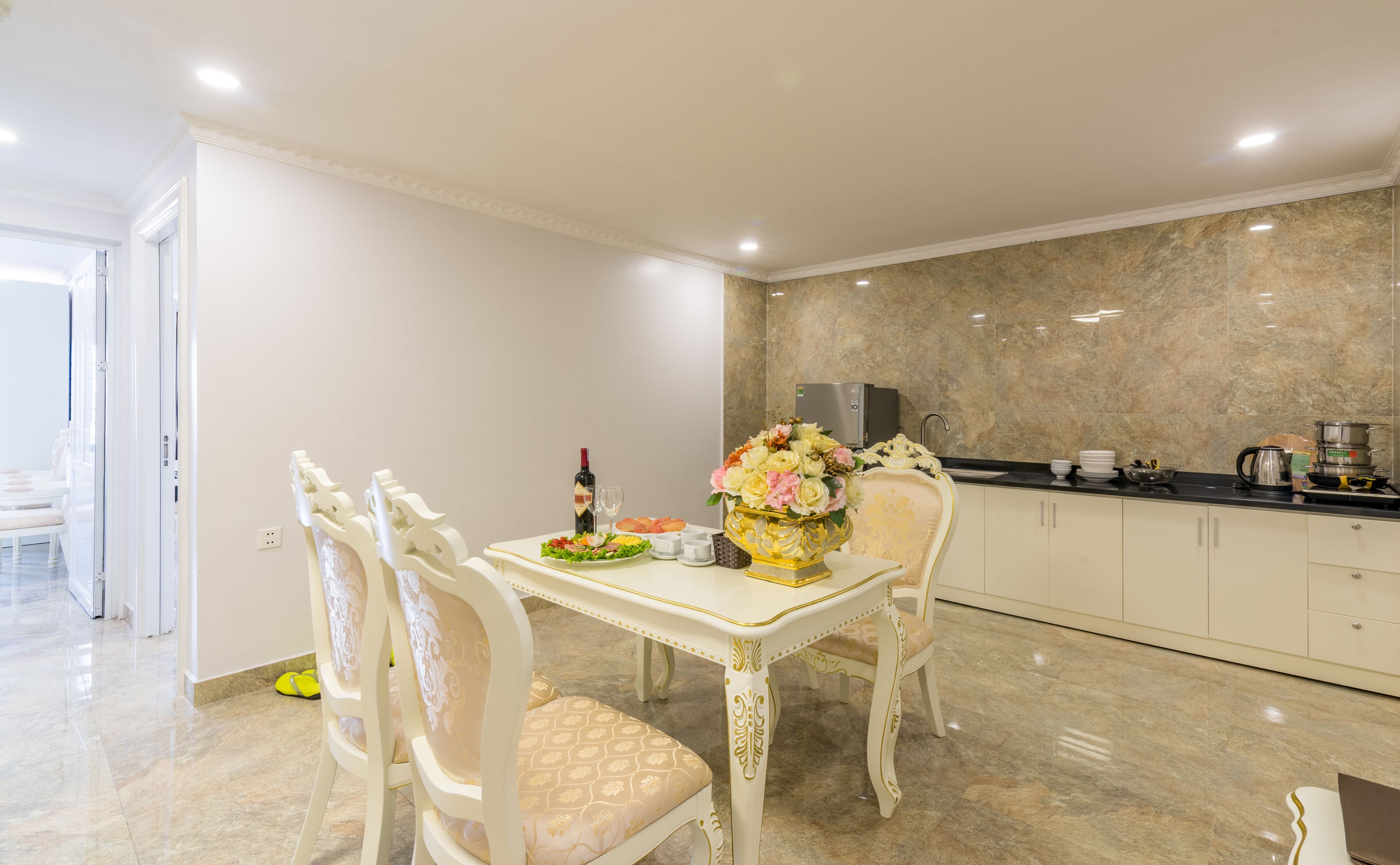 Un comedor con mesa y sillas blancas, un jarrón de flores y una cocina con salpicadero y armarios de mármol.