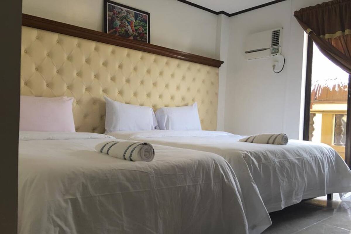 A hotel room with a large bed, a headboard, a wall-mounted air conditioning unit, and a window with curtains.