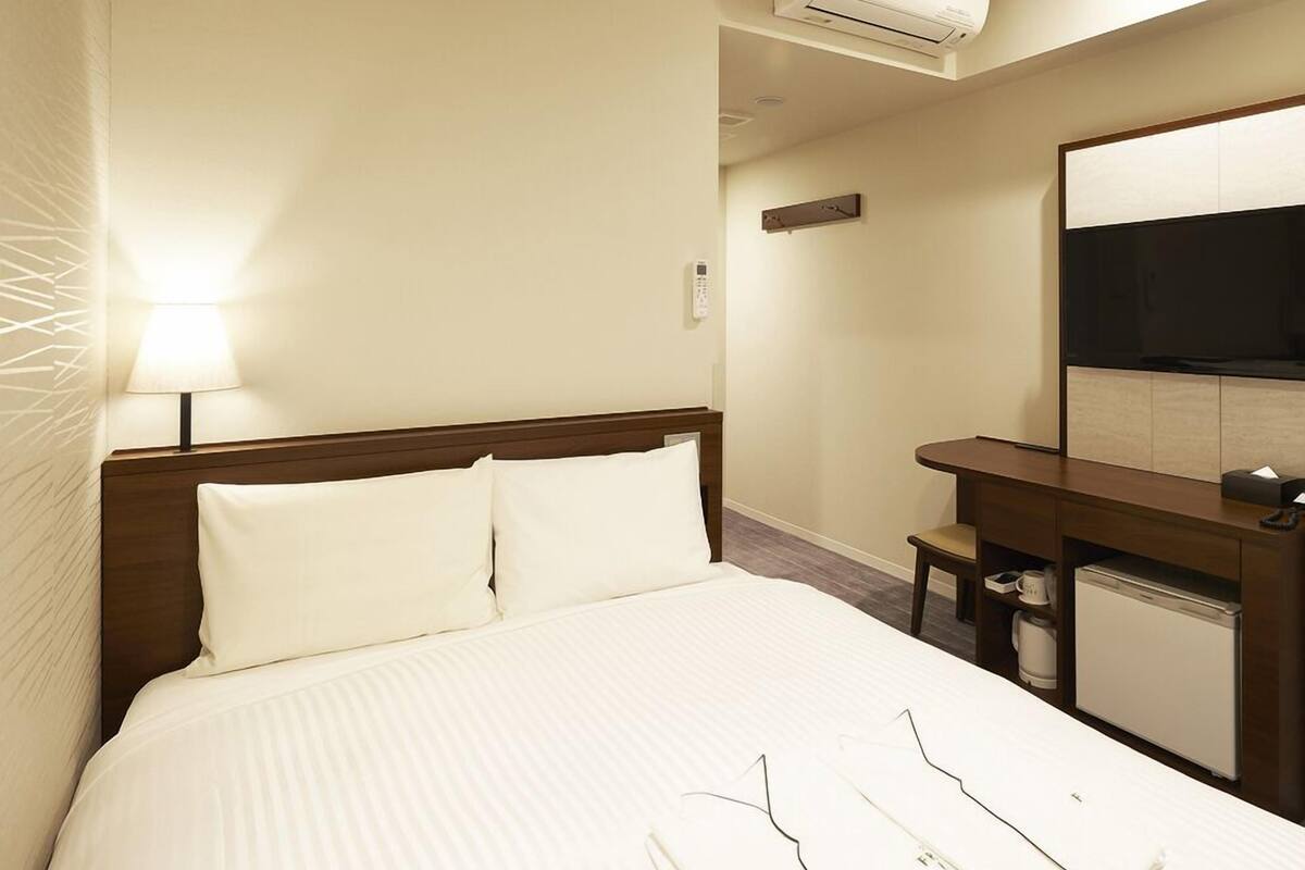 A hotel room with a bed, bedside lamp, air conditioning unit, desk, chair, and a television.
