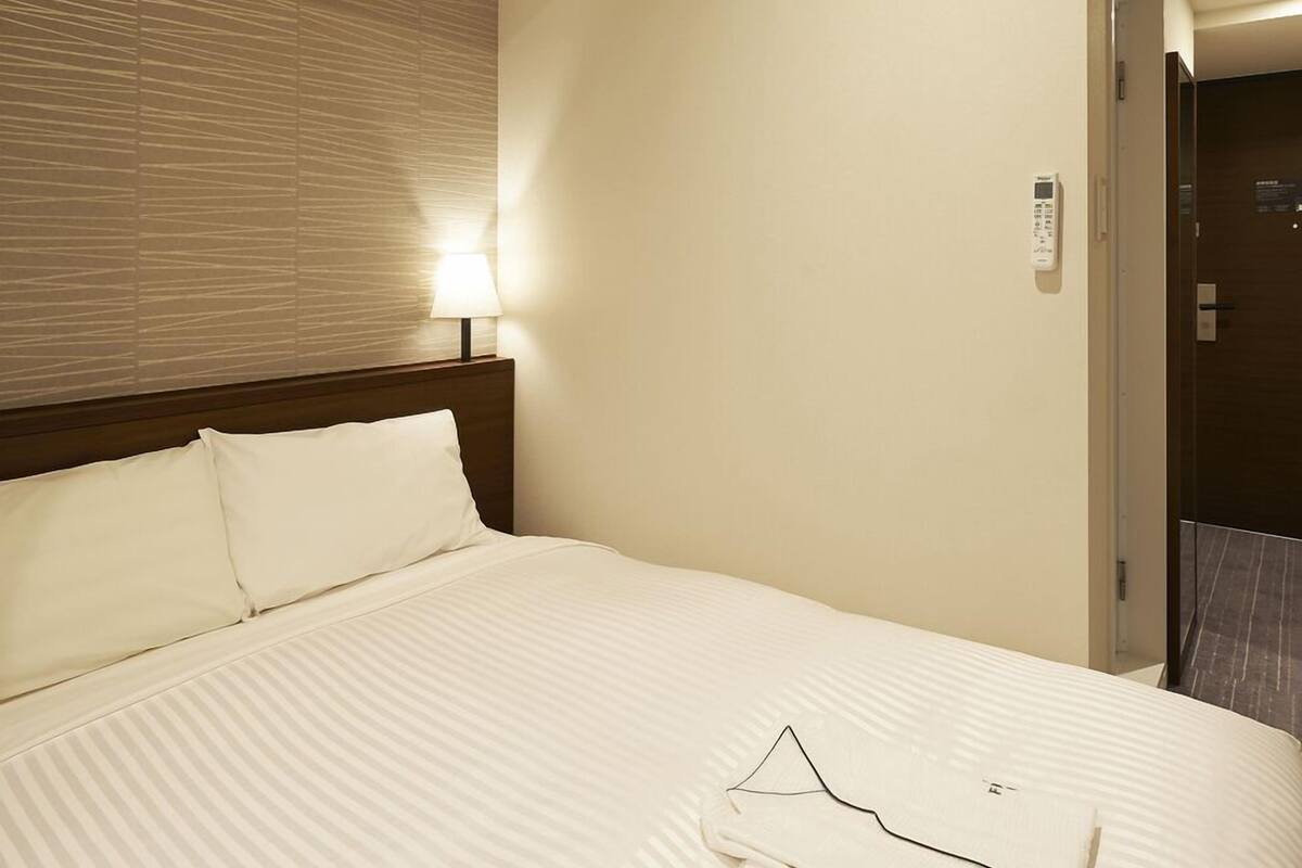A hotel room with a bed, a nightstand with a lamp, a wall-mounted air conditioner, and a door leading to another room.