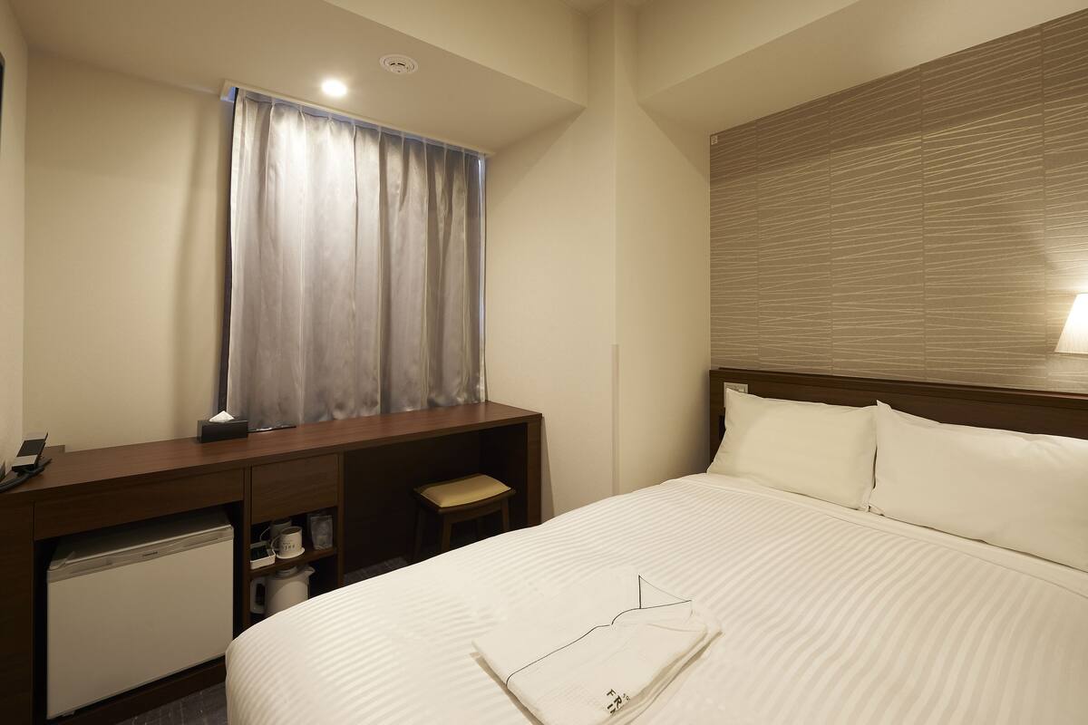 A hotel room with a bed, a desk, a chair, a small table, a lamp, and a window with curtains.