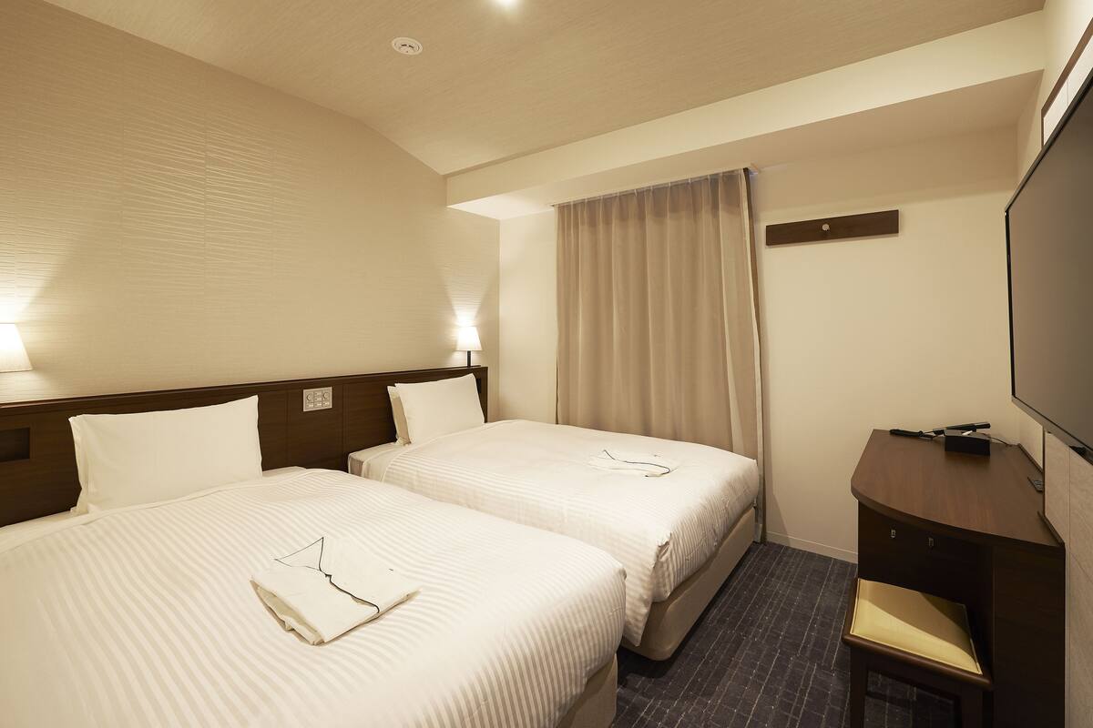 A hotel room with two beds, a desk, a chair, a television, and a window with curtains.