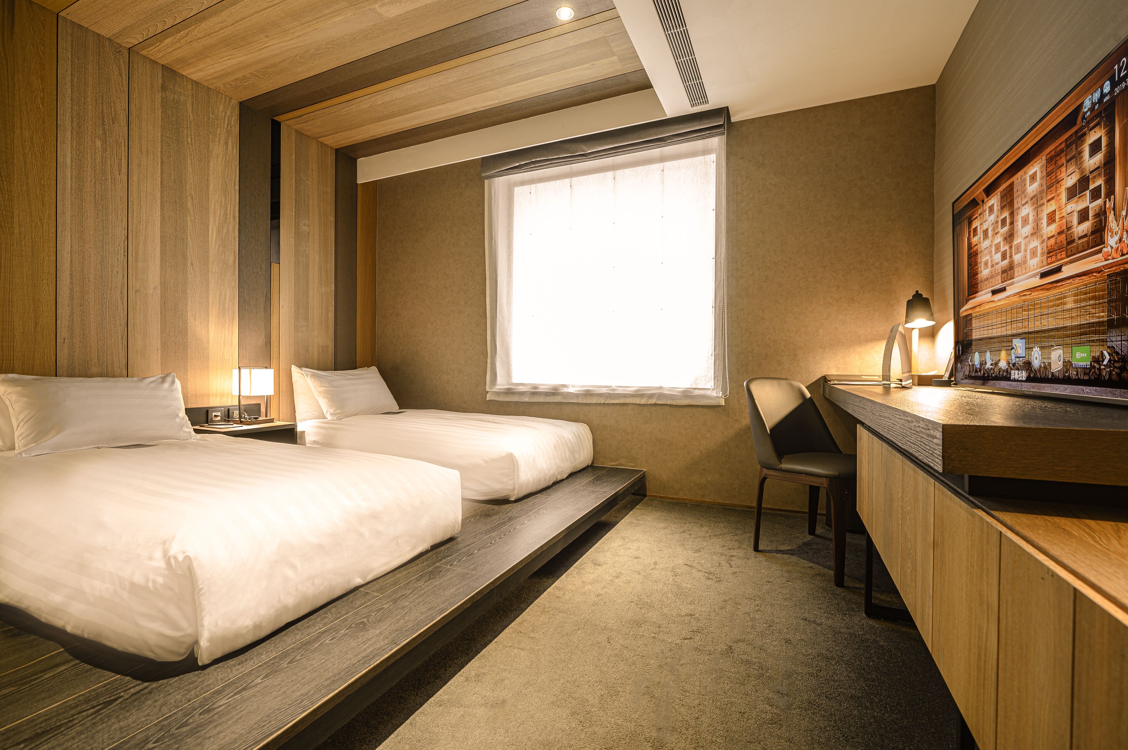 A hotel room with two beds, a desk with a lamp, a window, and a minibar.