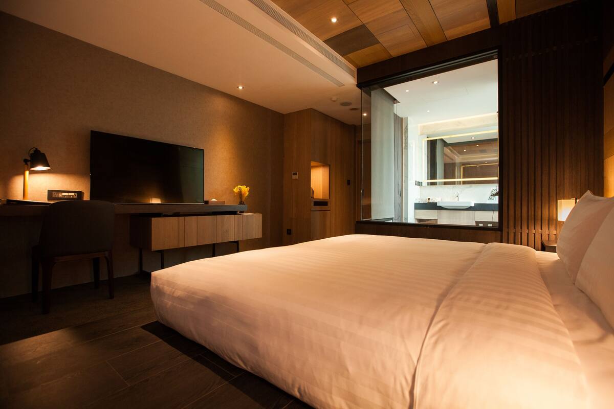 A hotel room with a large bed, a desk with a flat-screen TV, a chair, and a large window offering a view of the interior.