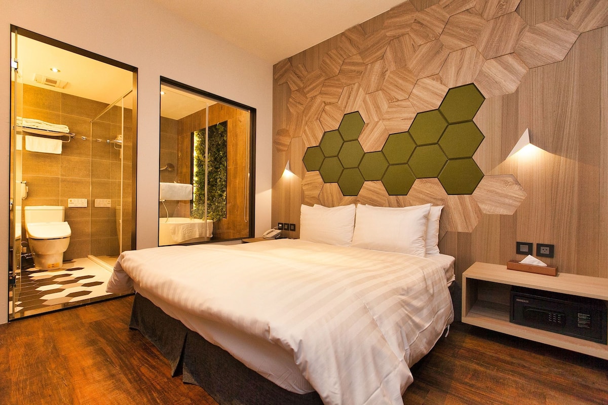 A modern hotel room with a large bed, a wooden wall with hexagonal panels, a bedside table with a lamp, and a mirror.
