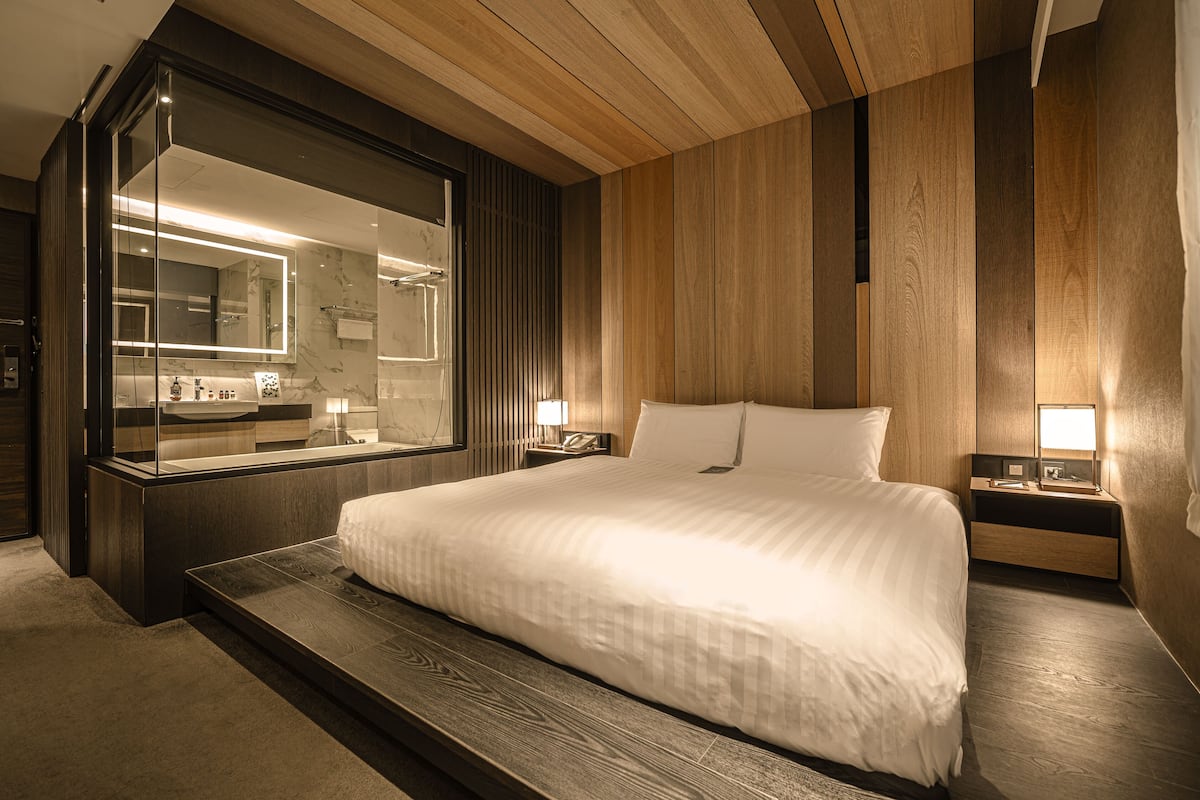 A modern hotel room with a large bed, wooden panel walls, a large mirror, and bedside lamps.