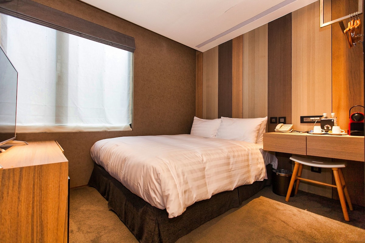 A hotel room with a bed, a desk, a chair, a window, and a wall with vertical wooden panels.