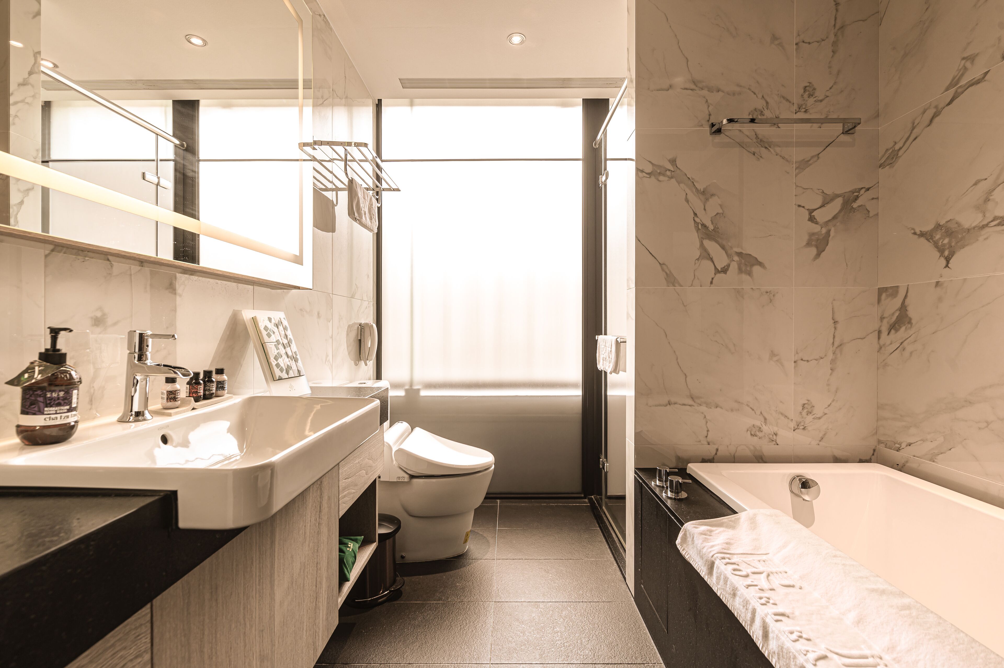 Een moderne badkamer met een marmeren wand, een bad, een toilet en een wastafel met toiletartikelen.