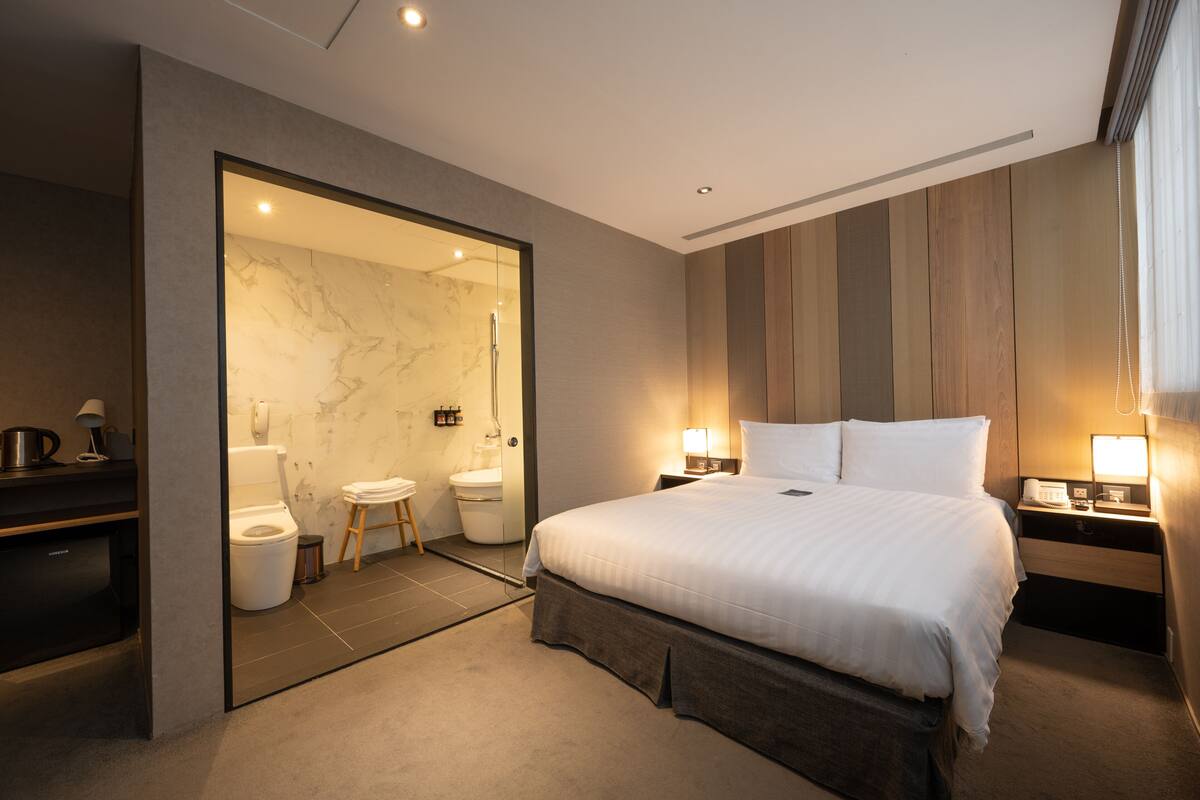 A modern hotel room with a large bed, bedside lamps, a wooden panel wall, and a bathroom with a marble shower and toilet.