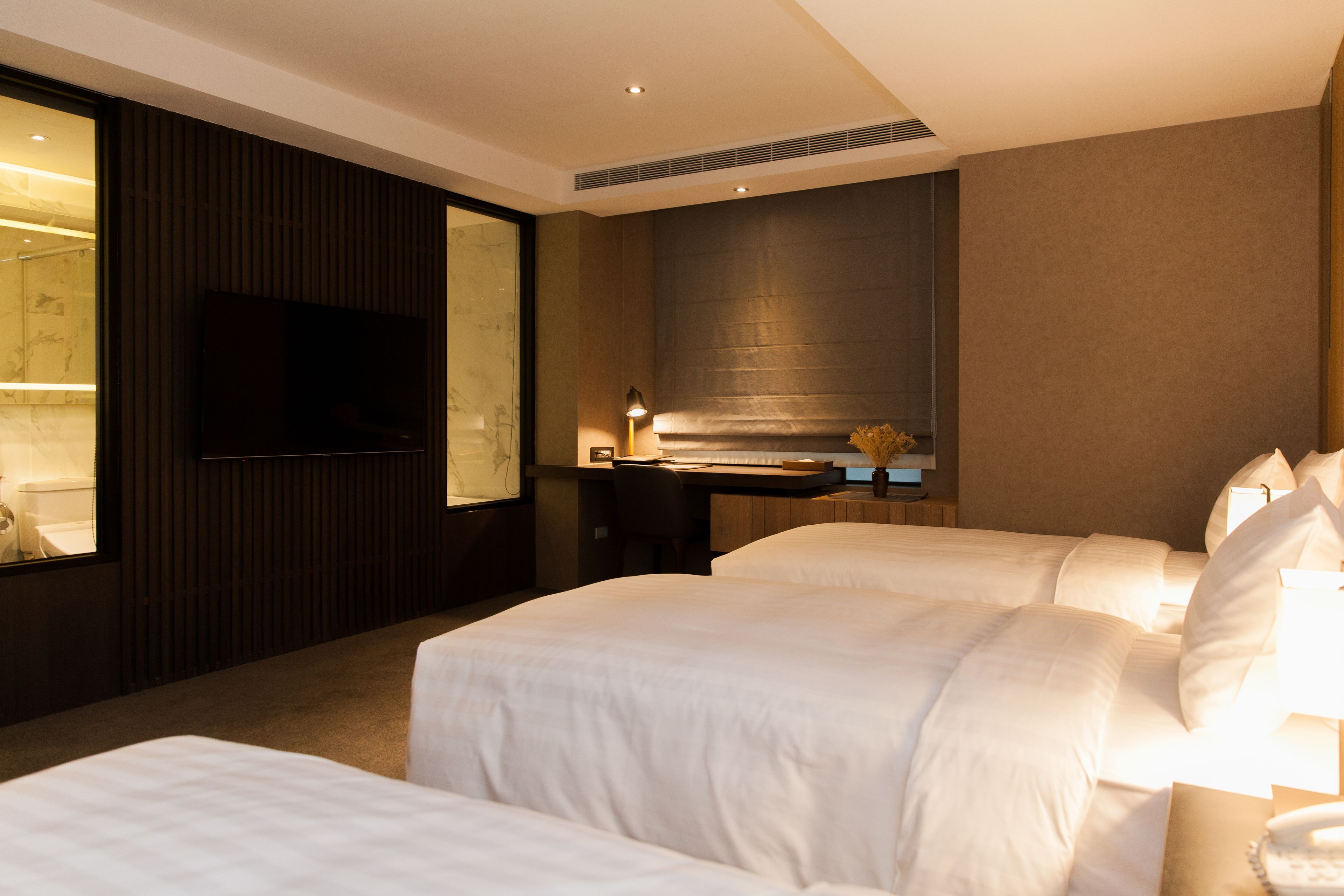 A hotel room with two beds, a desk, a flat-screen TV, and a bathroom with a shower.