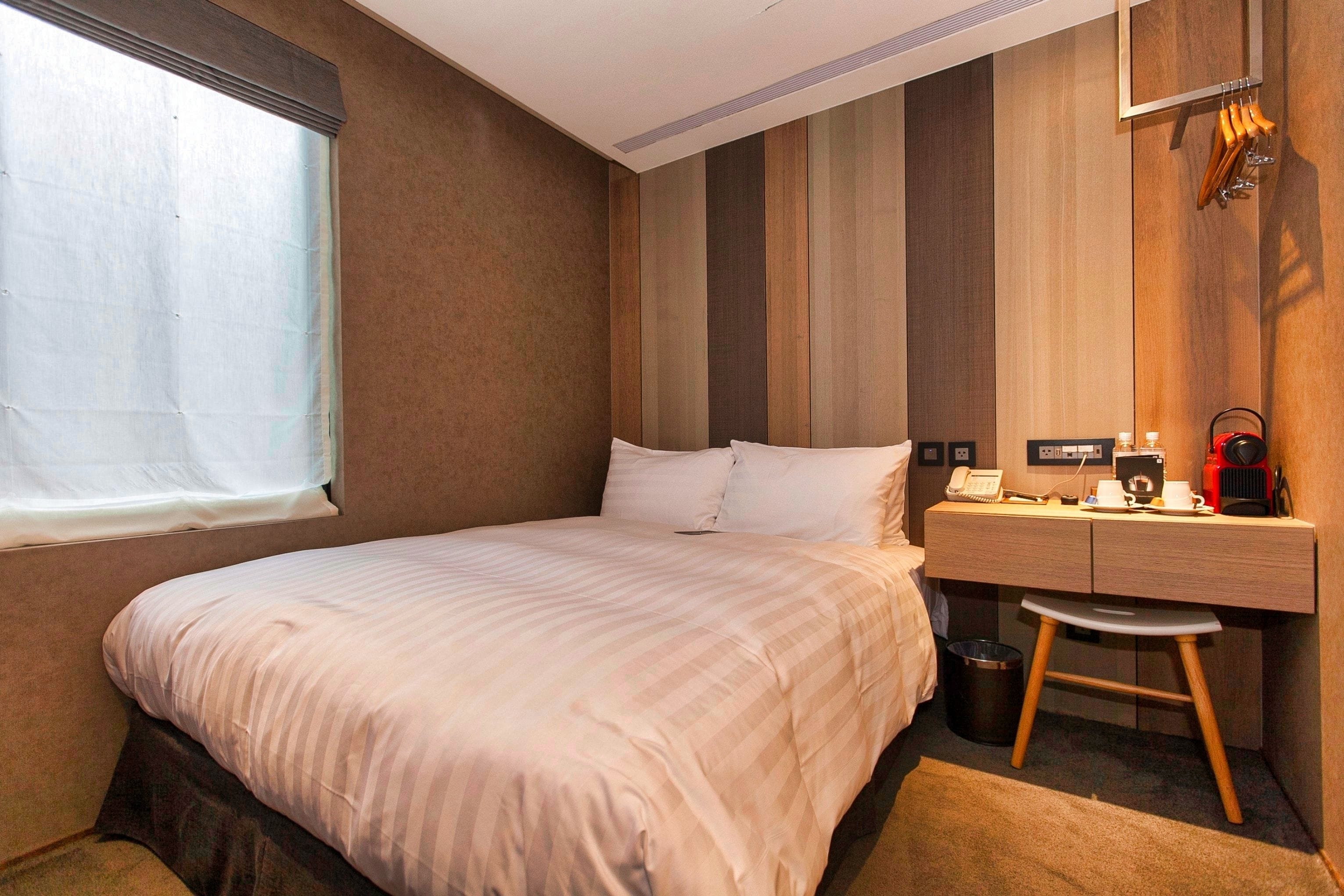Habitación de hotel con cama, escritorio, silla, cesto de basura y ventana con persianas.