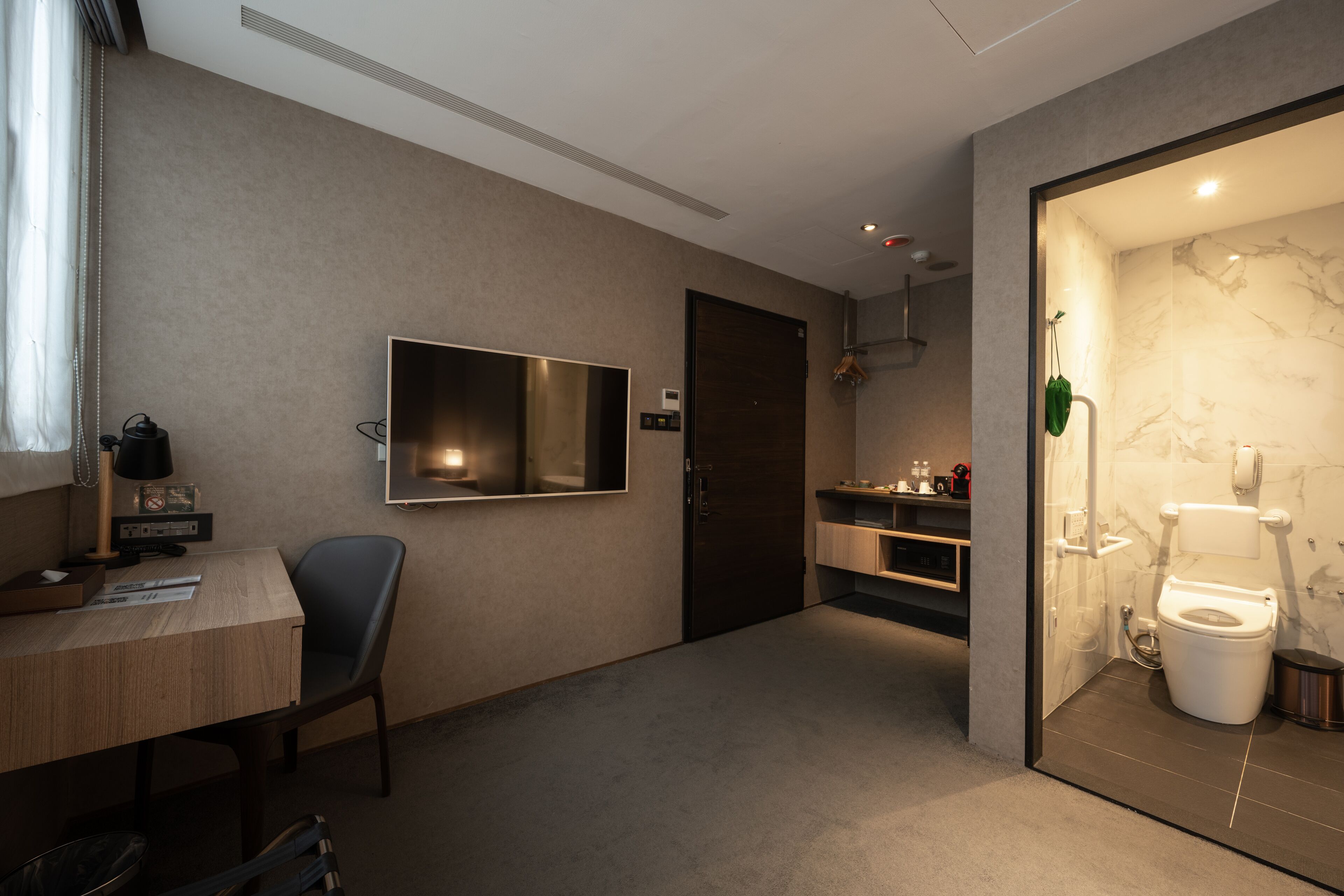 A modern hotel room with a desk, chair, TV, and a bathroom with a toilet and sink.