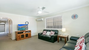 A living room with a green sofa, a wooden entertainment unit, a ceiling fan, and a window with blinds.