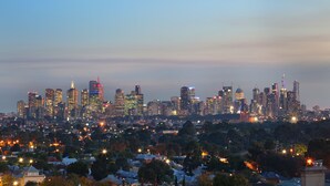 A city skyline at dusk with illuminated buildings and a dense residential area below. - StayCentral - Northcote Hill Penthouse (Northcote)