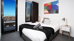 A hotel room with a bed, a nightstand, a lamp, a window with curtains, and a colorful abstract painting on the wall. - StayCentral - Northcote Hill Penthouse (Northcote)