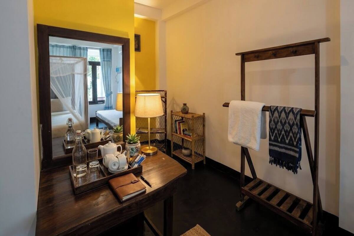 Una habitación con un escritorio de madera, un espejo, una mesita de noche con lámpara, una estantería con libros y un perchero para toallas.