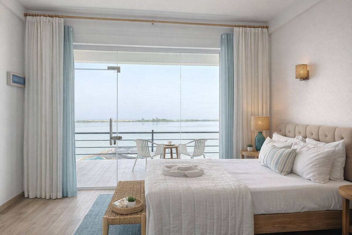 Habitación de hotel con una cama grande, una pequeña mesa con sillas, vista al mar y un balcón con cortinas.