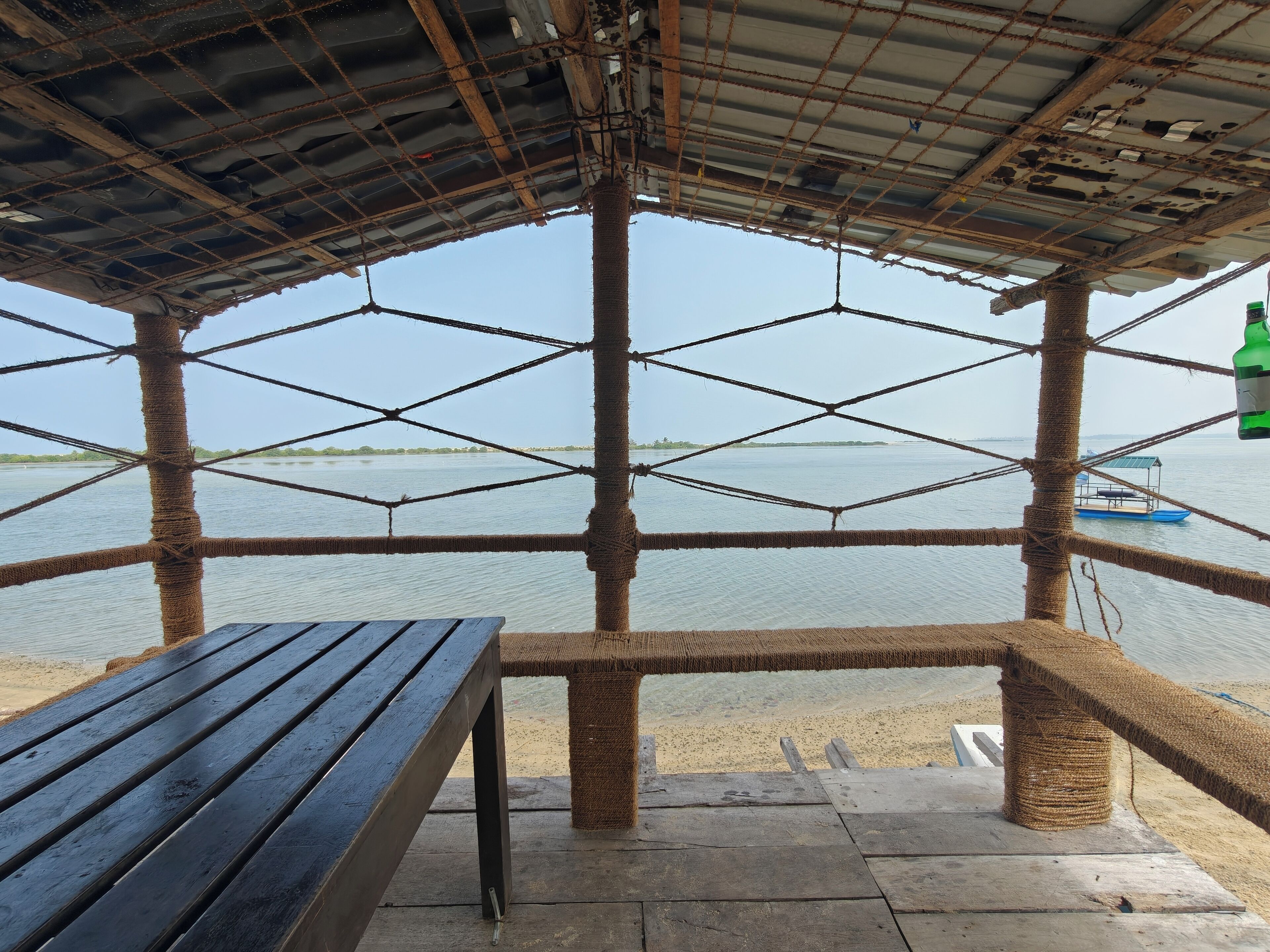 A wooden bench and a rope-wrapped structure overlooking a calm body of water with a boat in the distance.