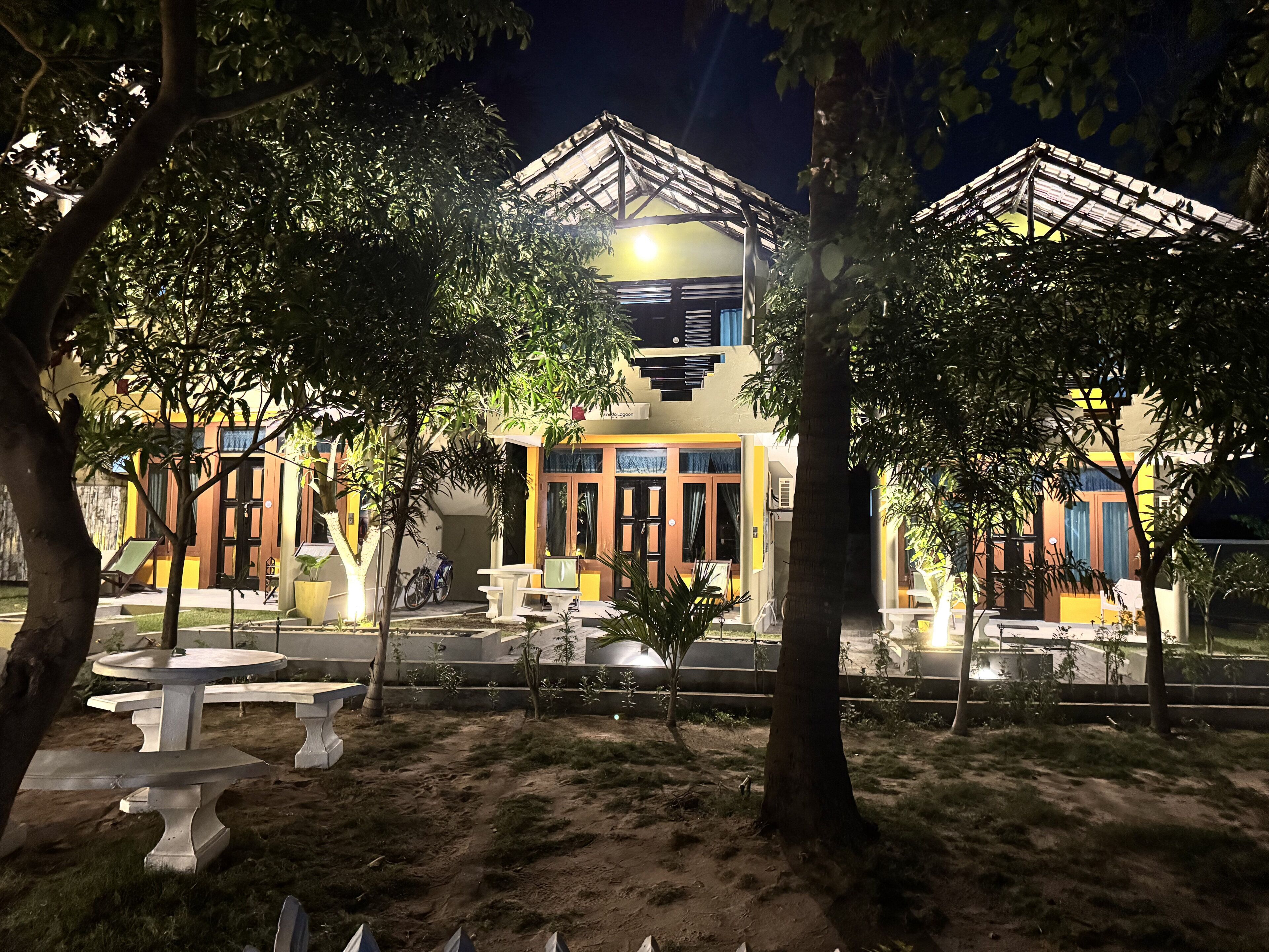 A building with a covered outdoor seating area, surrounded by trees, and a paved walkway.