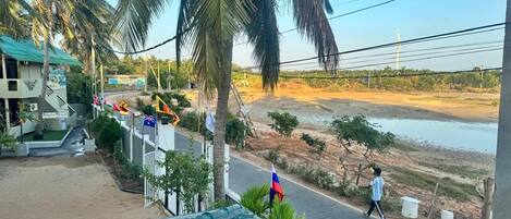 Une vue depuis un balcon donnant sur une route bordée de palmiers, une rivière et un bâtiment avec un toit bleu.