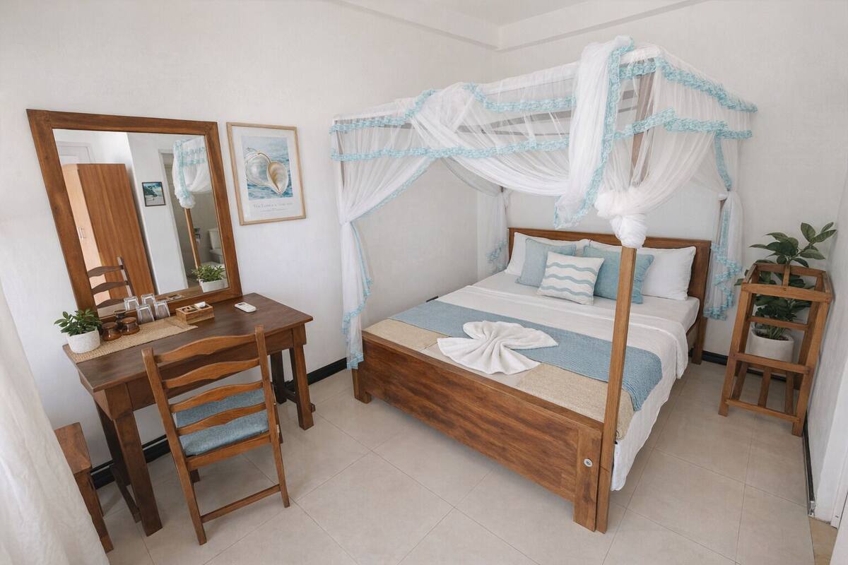 Un dormitorio con cama de madera, dosel, escritorio con sillas, espejo y mesita de noche con una planta.