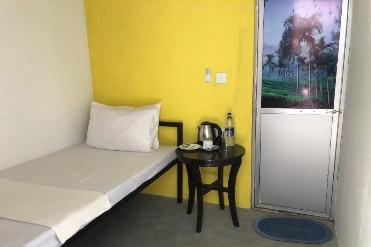 Una habitación pequeña con cama, una mesa con hervidor y botella de agua, una ventana con vista a palmeras y una puerta.