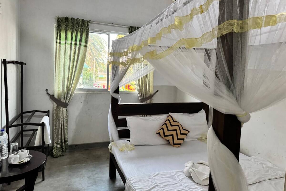 Una habitación con cama, una mesita con silla, una ventana con cortinas y vistas al exterior.