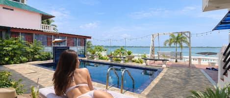 A pool area with a view of the sea, a lounge chair, and a table with a drink.
