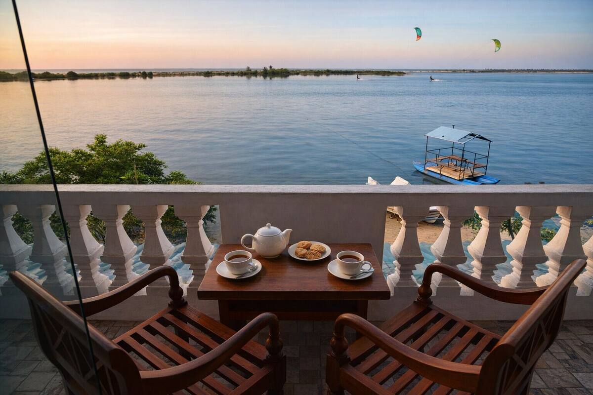 Un balcón con una mesa lista para dos personas, con vista a una masa de agua tranquila con barcos y cometas a lo lejos.