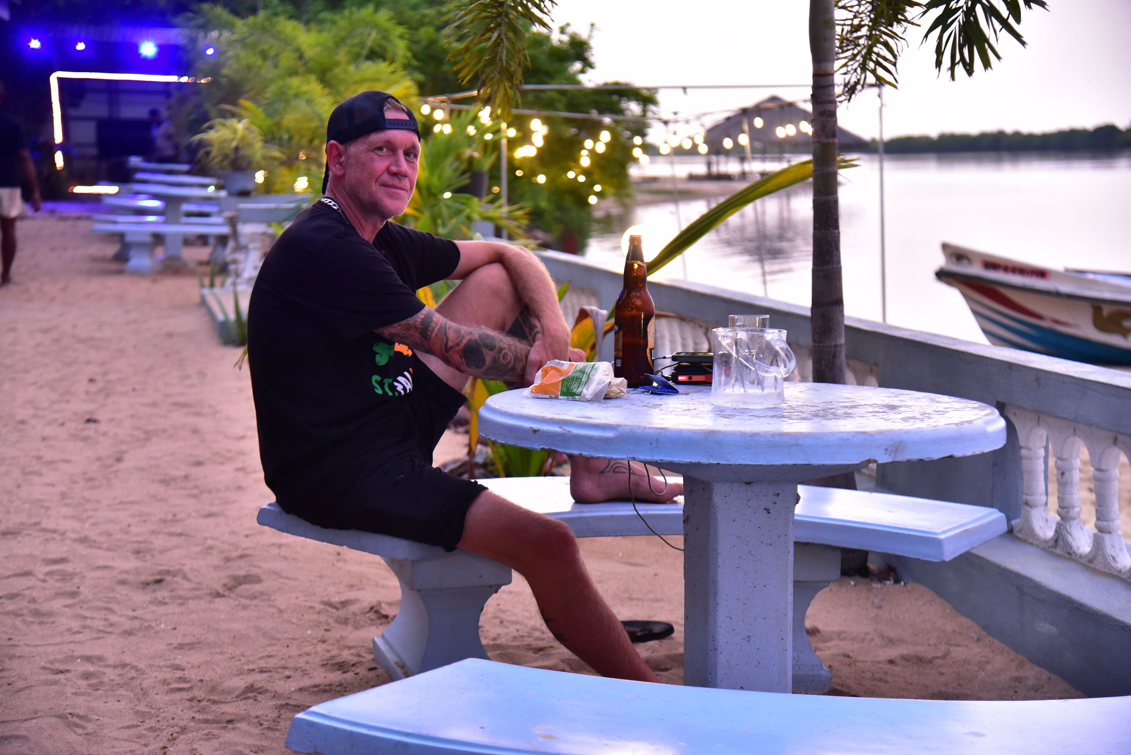 Un homme est assis à une table, devant une bouteille et des verres, et il surplombe une étendue d'eau calme, avec un bateau visible au loin.