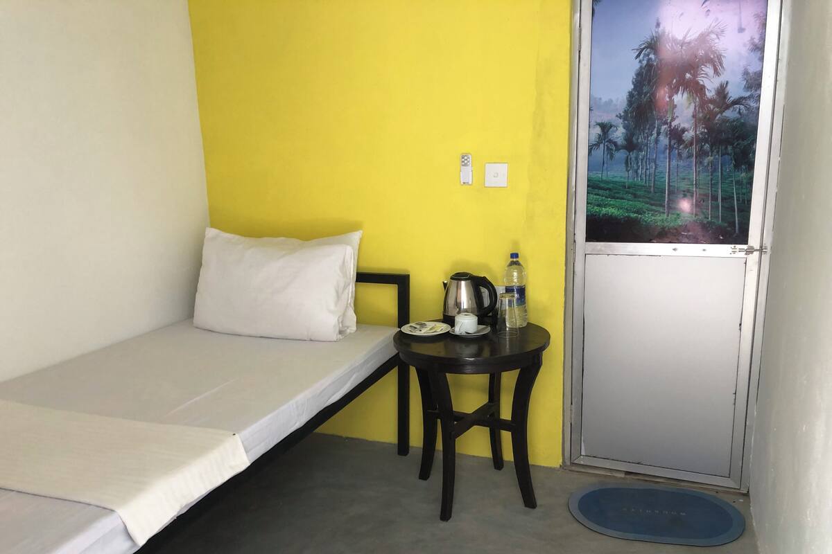 Una cama individual con una almohada blanca, una mesita con un hervidor, una botella y una taza, y vistas a palmeras a través de una puerta.