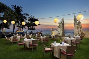 Un espace de restauration en plein air, avec des tables et des chaises, des guirlandes lumineuses et une vue sur l’océan au coucher du soleil.