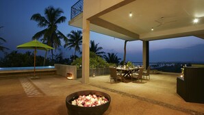 Un área de comedor al aire libre con una mesa para dos, un hogar para fuego con flores y vista al océano al atardecer.