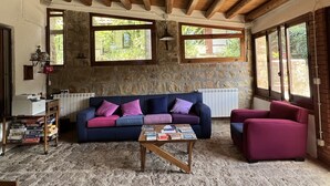 Un salon chaleureux avec un mur en pierre, des poutres apparentes en bois au plafond, un canapé bleu agrémenté de coussins violets, une table basse en bois et un fauteuil rouge.