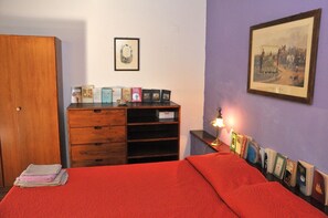 Un dormitorio con cama, un armario de madera, una estantería con libros y fotografías enmarcadas, y un cuadro colgado en la pared.