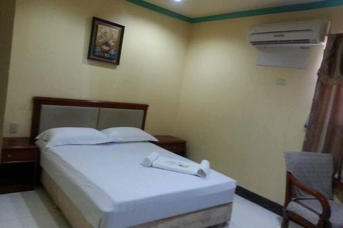 A hotel room with a bed, a nightstand, a chair, an air conditioning unit, and a painting on the wall.