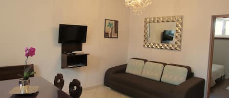 Ein Wohnzimmer mit braunem Sofa, einem an der Wand montierten Fernseher, einer Kristallleuchtre und einem Esstisch mit Stühlen.