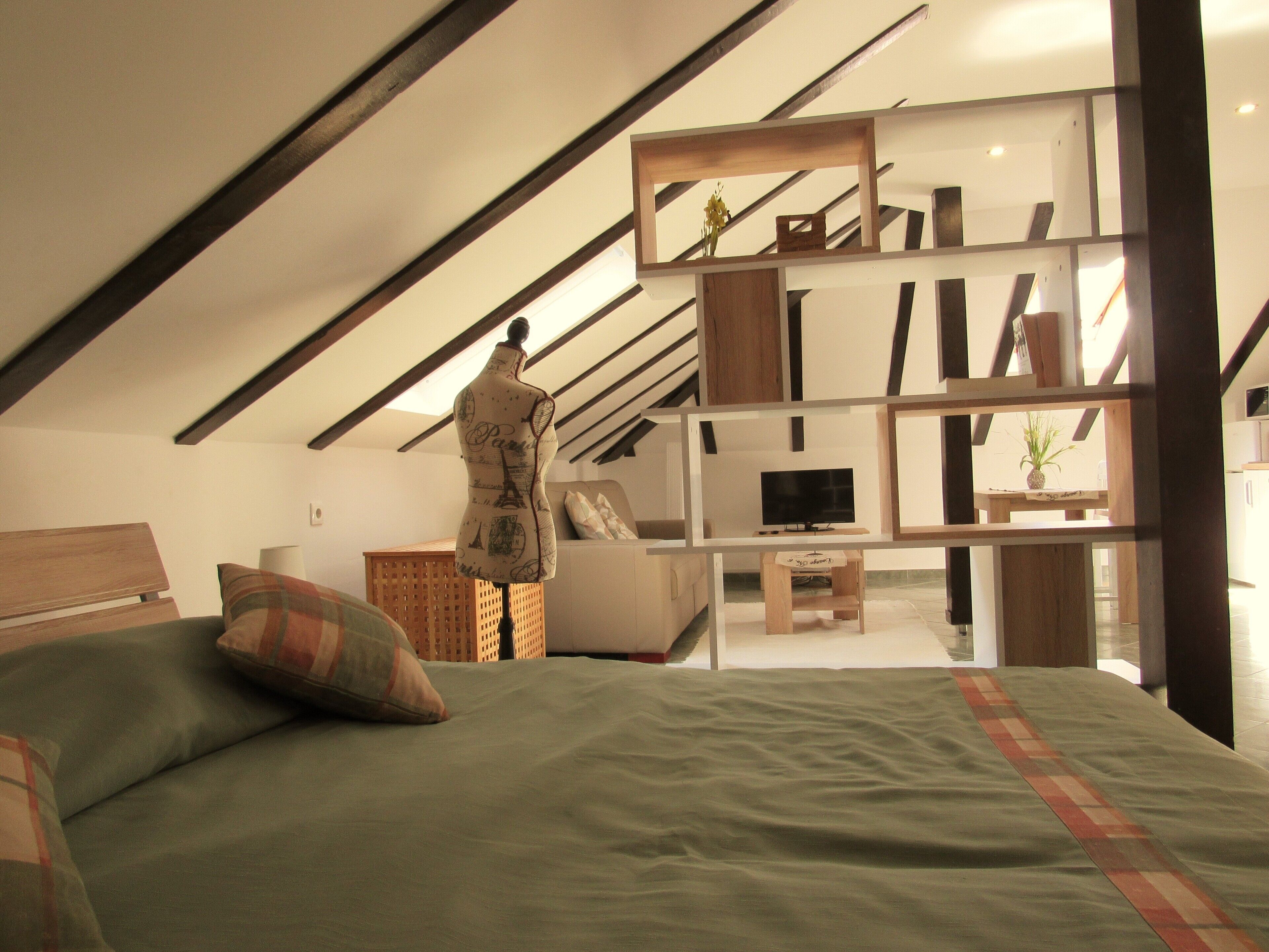 A bedroom with a sloped ceiling, a bed with green bedding, a mannequin, a TV on a stand, and a wooden shelf unit.