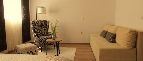 Habitación de hotel con cama, un sillón estampado de cebra, una pequeña mesa redonda con una planta y un sofá beige con cojines decorados.