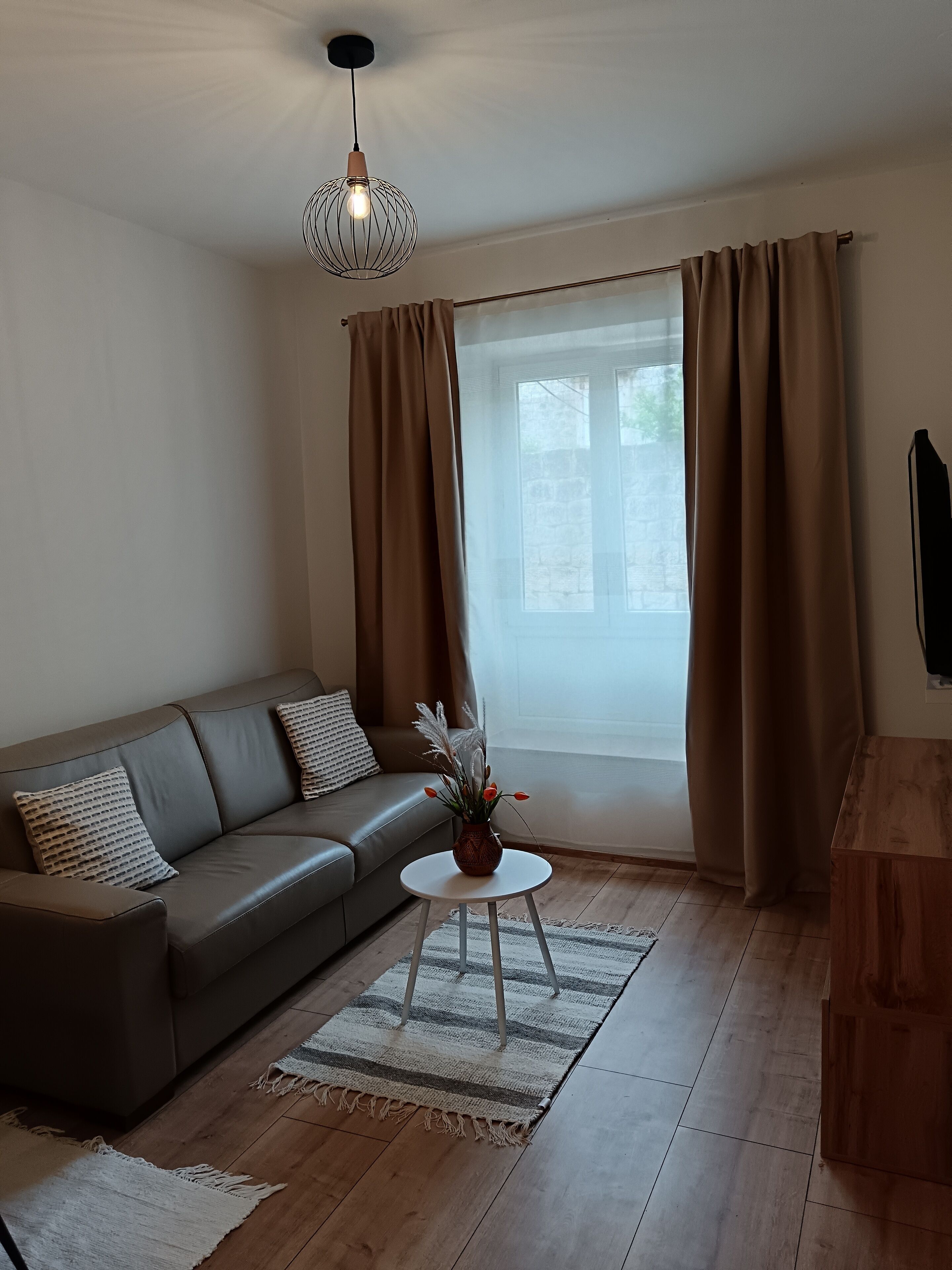 Un salon moderne avec un canapé gris, une petite table ronde ornée d’un vase, un meuble en bois et une grande fenêtre avec des rideaux.