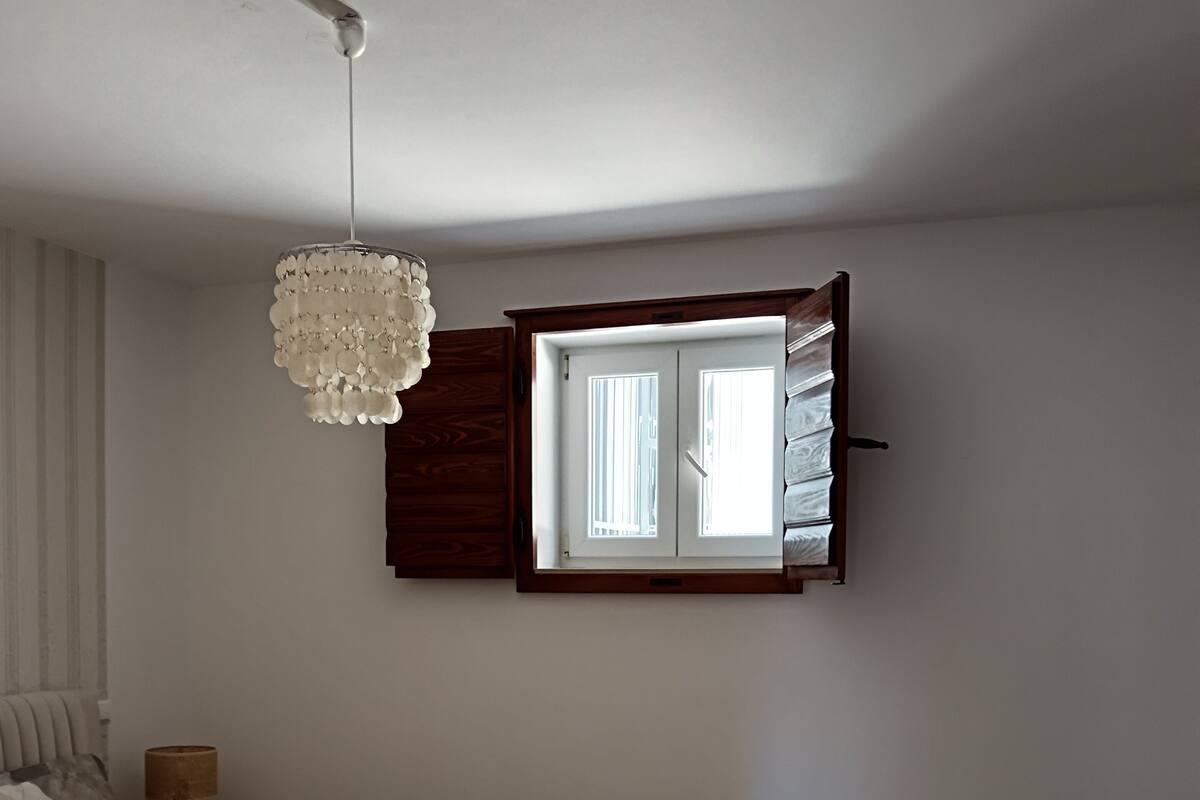 A bedroom with a bed, a chair, a small table, a window with curtains, and a chandelier.