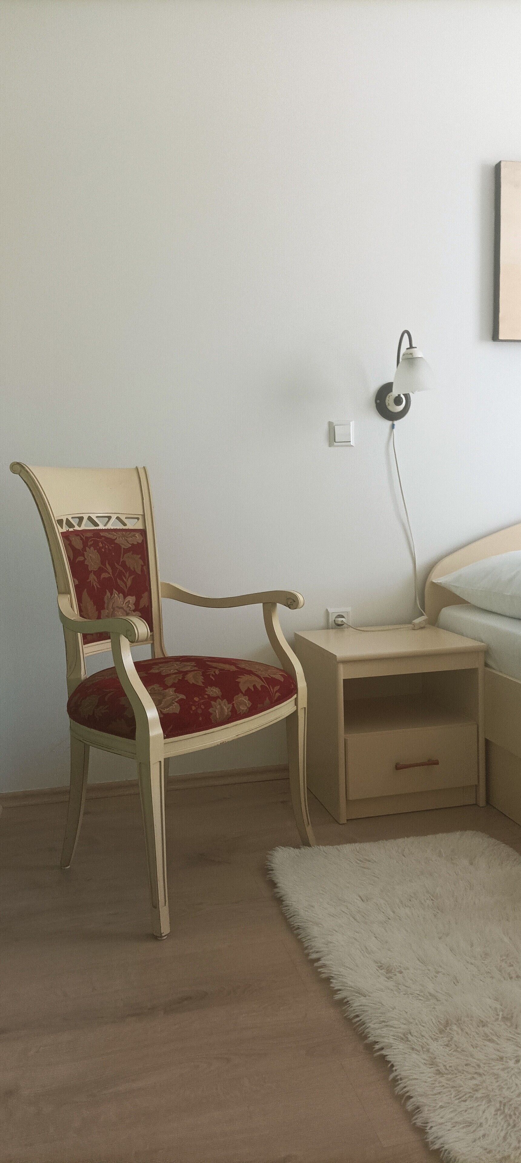 A room with a wooden chair with a floral cushion, a bedside table with a lamp, and a white fluffy rug.