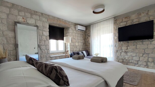 A bedroom with a stone wall, a bed with white bedding, a bedside lamp, a TV mounted on the wall, and a window with curtains. - House Klaudija (Trogir)