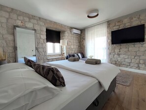 A bedroom with a stone wall, a bed with white bedding, a bedside lamp, a TV mounted on the wall, and a window with curtains. - House Klaudija (Trogir)