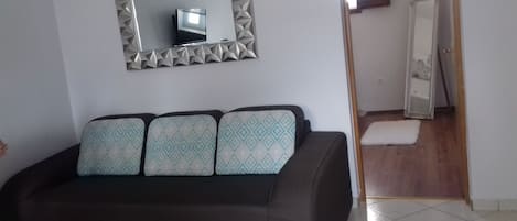 Una habitación con un sofá oscuro, un cojín estampado, un espejo con marco geométrico y suelo de madera.