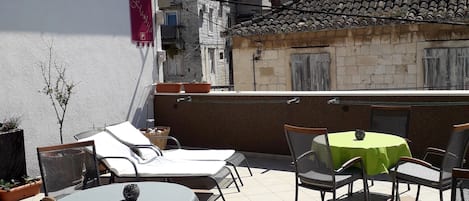 An outdoor patio area with round tables and chairs, a sign for rooms and apartments, and a view of traditional buildings with tiled roofs.