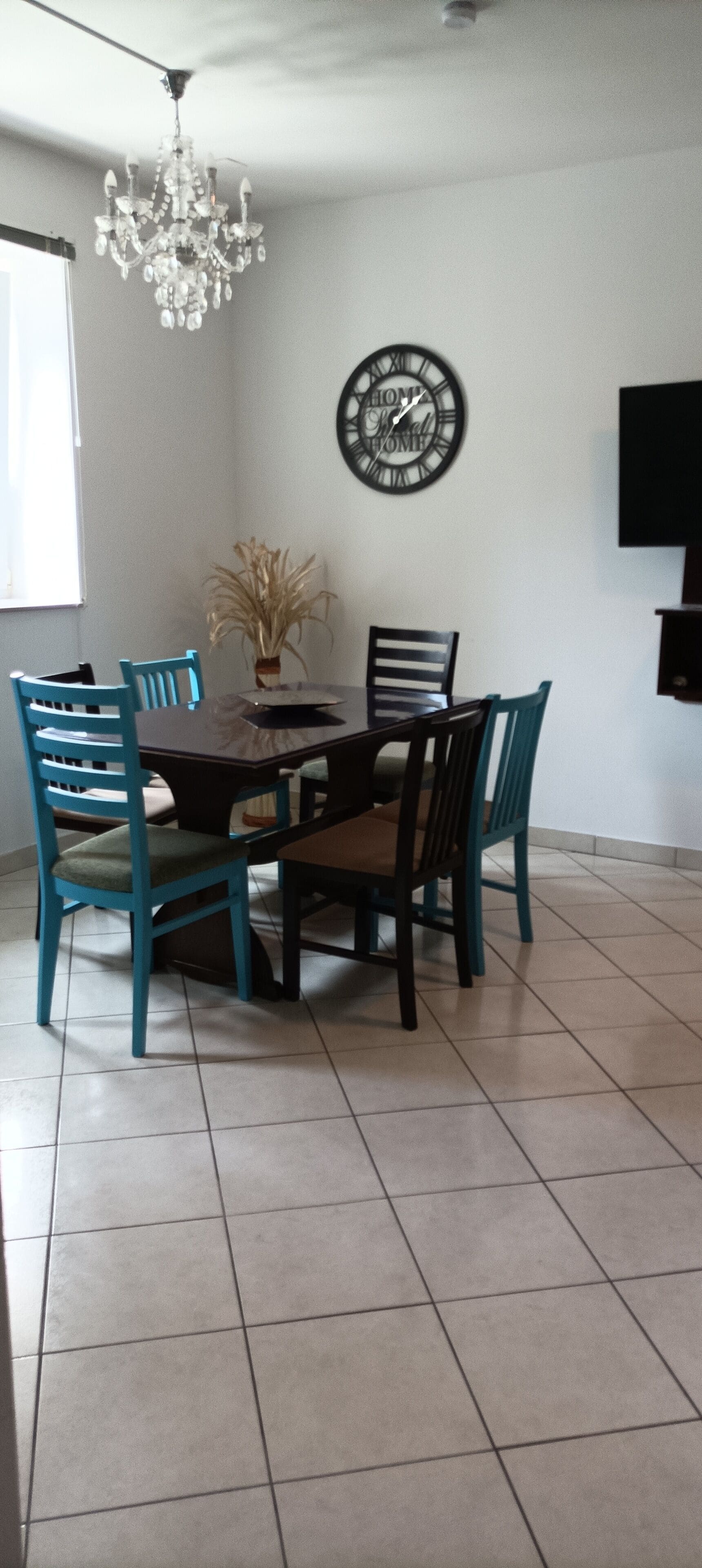 A dining area with a round table, four chairs, a chandelier, a wall clock, and a flat-screen TV.