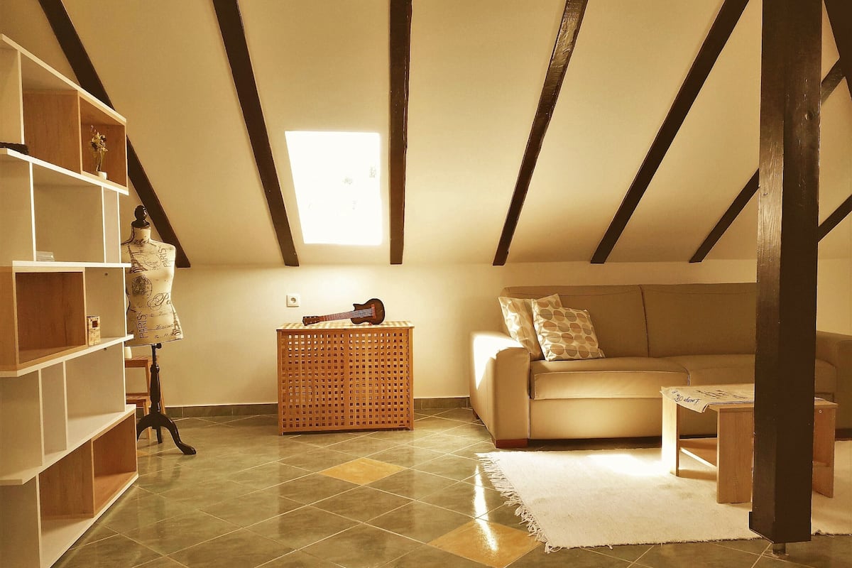 A cozy attic room with exposed wooden beams, a tiled floor, a sofa, a small table, and a mannequin.