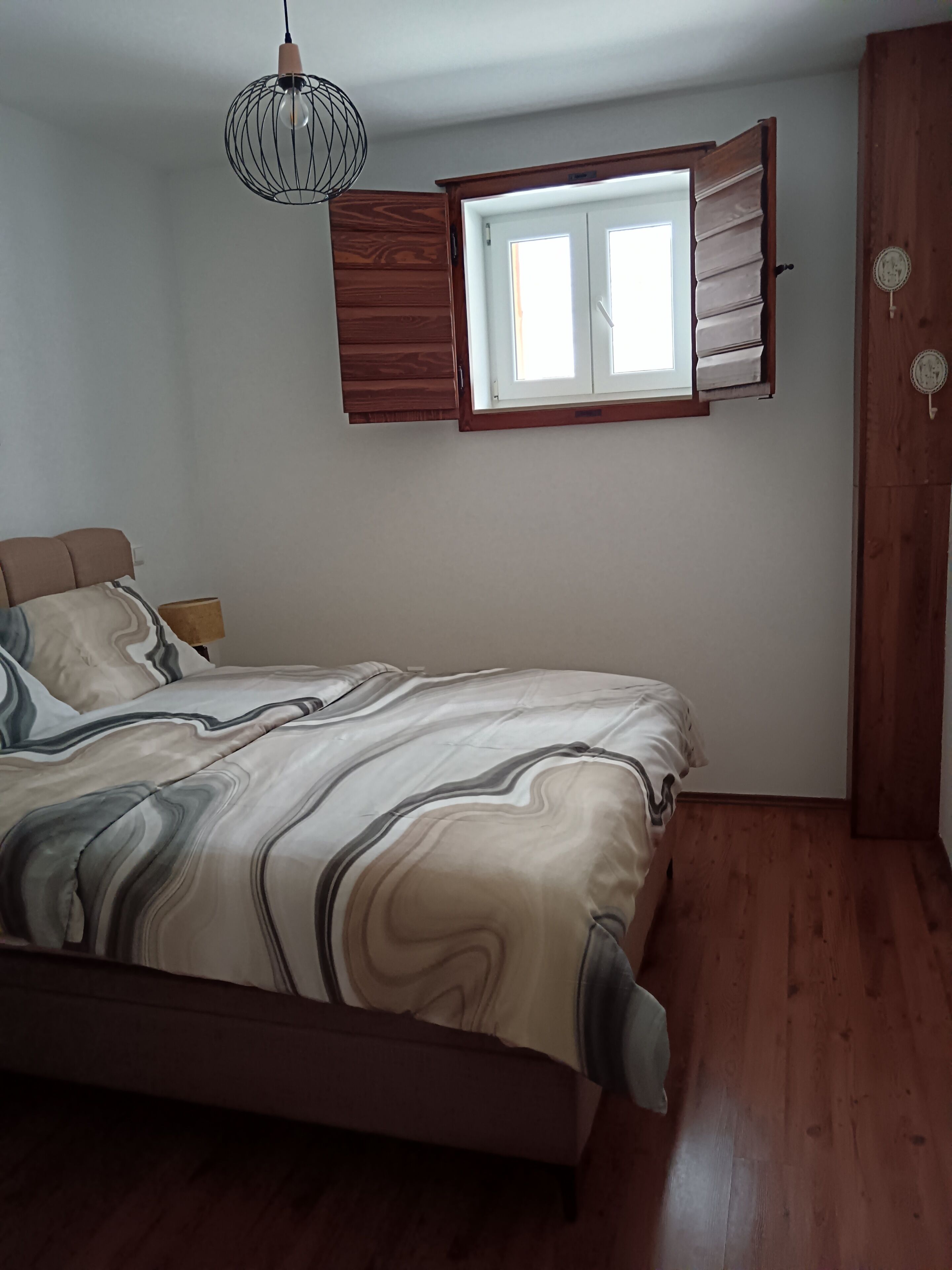 Ein Schlafzimmer mit Bett, einem Fenster mit Rollläden, Holzboden und einer Pendelleuchte.