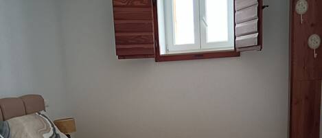 A bedroom with a bed, a window with shutters, a wooden floor, and a hanging light fixture.